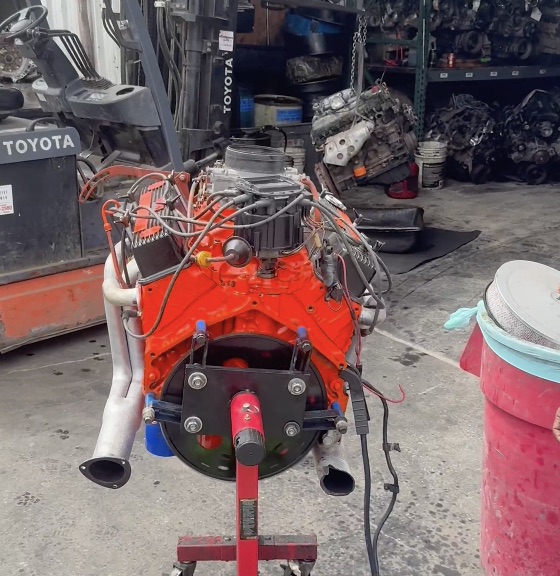 Rebuilt orange V8 engine on a stand at Villanueva Auto, with a forklift and used parts behind it